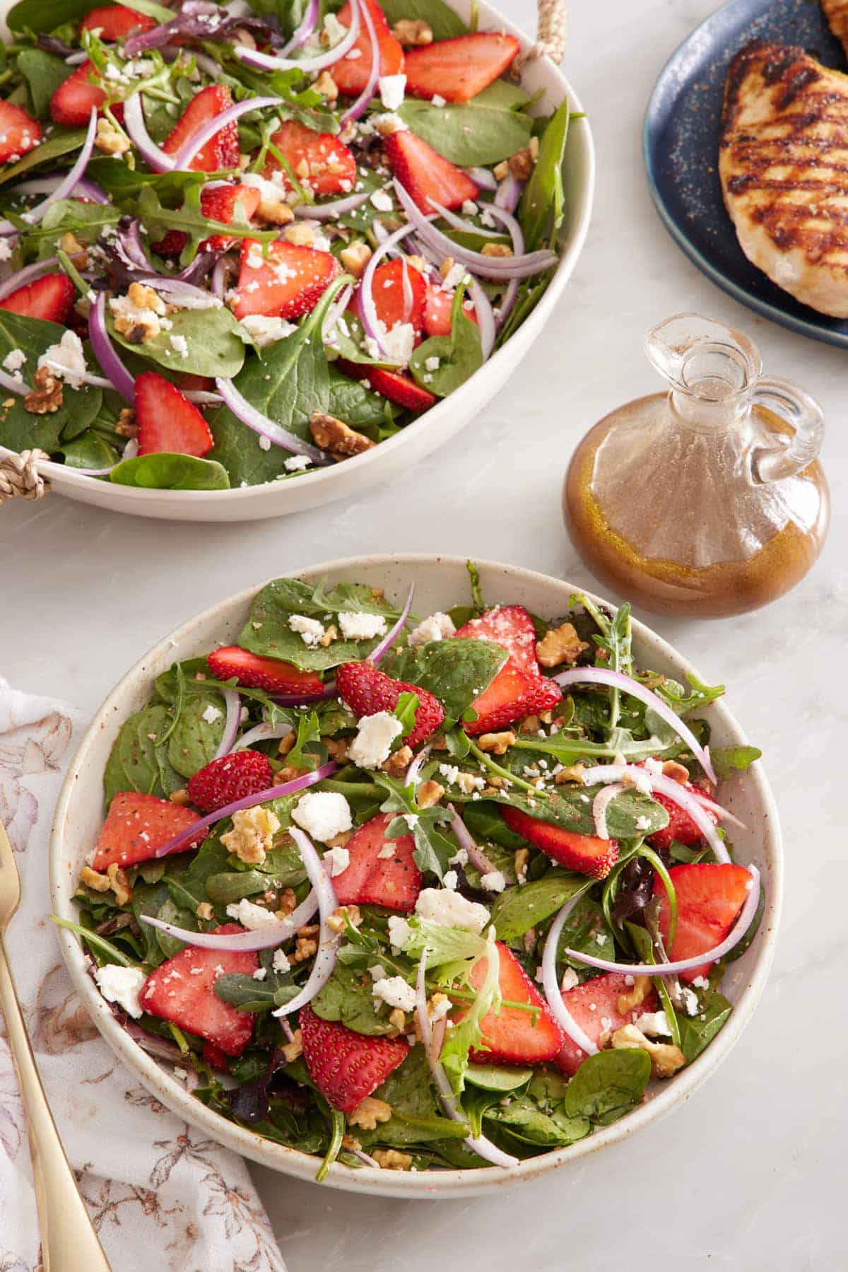 A bowl of strawberry salad with a platter more in the background along with a jar of salad dressing and a chicken breast.