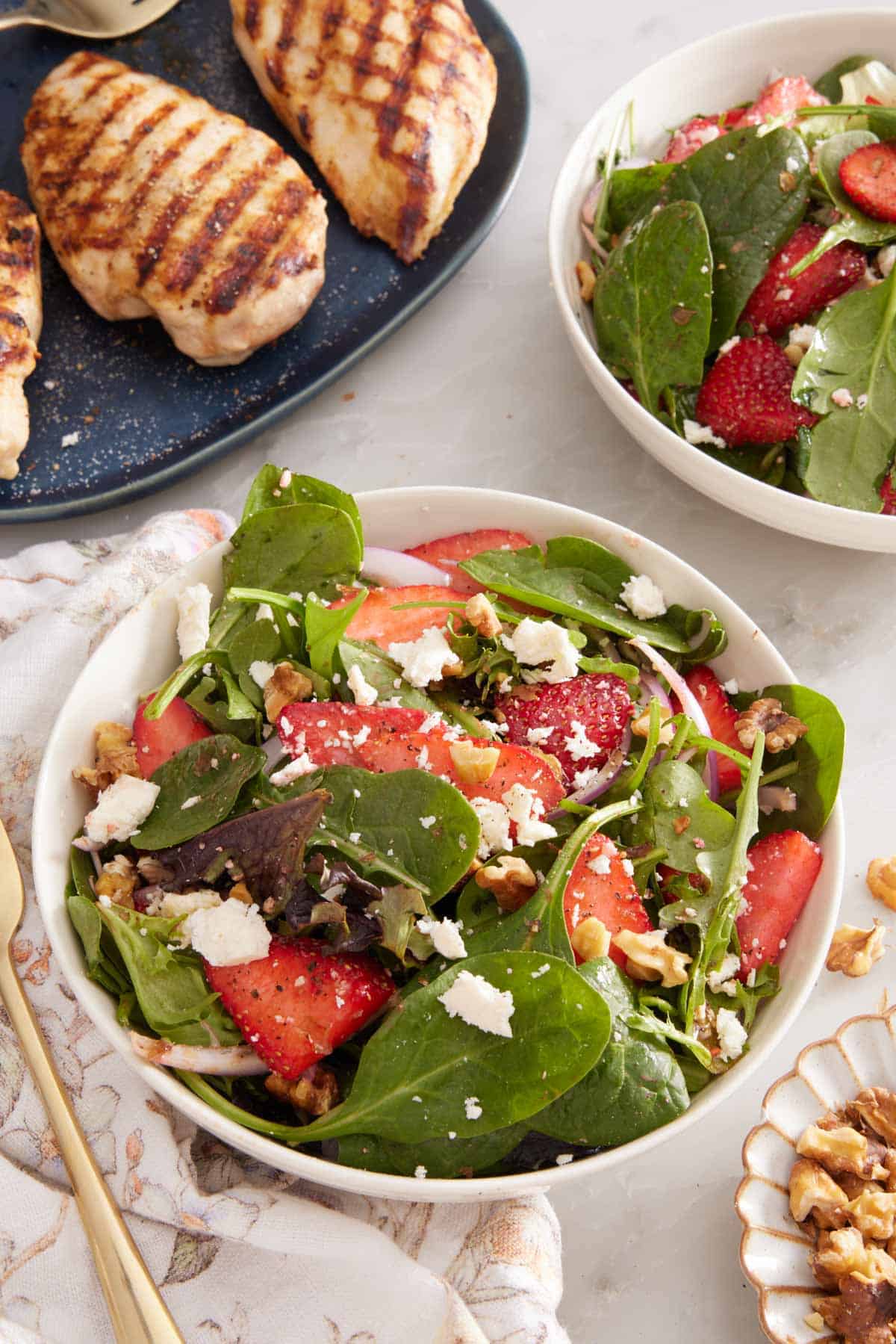 A bowl of strawberry salad with a second on off to the side with some grilled chicken breasts in the background.
