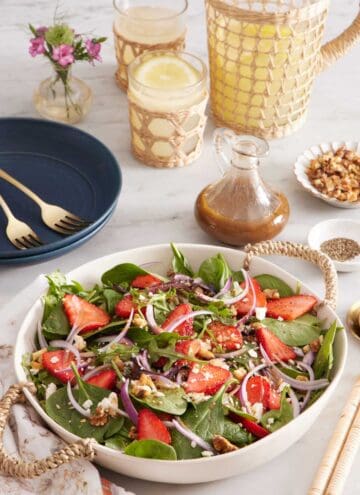 A large platter of strawberry salad. A pitcher of lemonade in the background with glasses, salad dressing, flowers, plates, and forks.