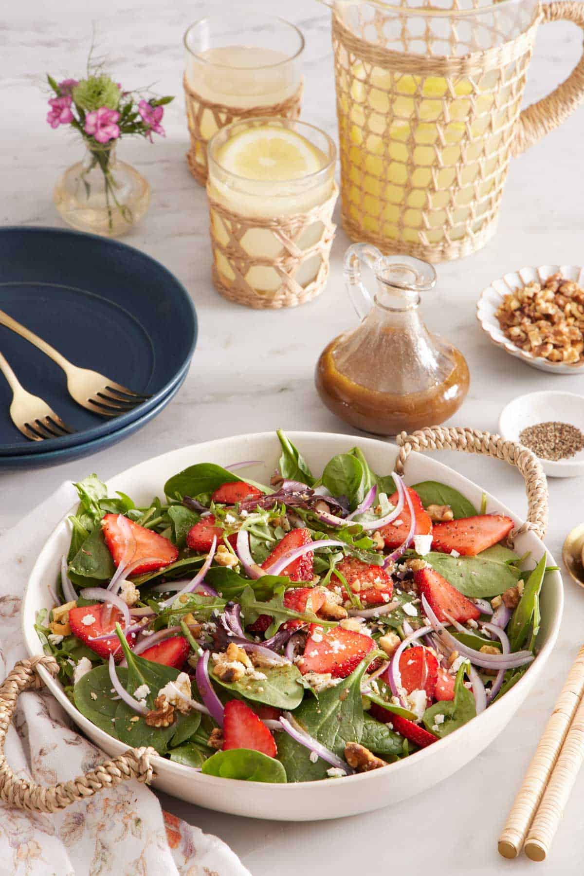 A large platter of strawberry salad. A pitcher of lemonade in the background with glasses, salad dressing, flowers, plates, and forks.