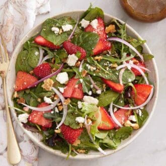 Overhead view of a plate of strawberry salad with a fork on the side along with chopped walnuts and flowers.