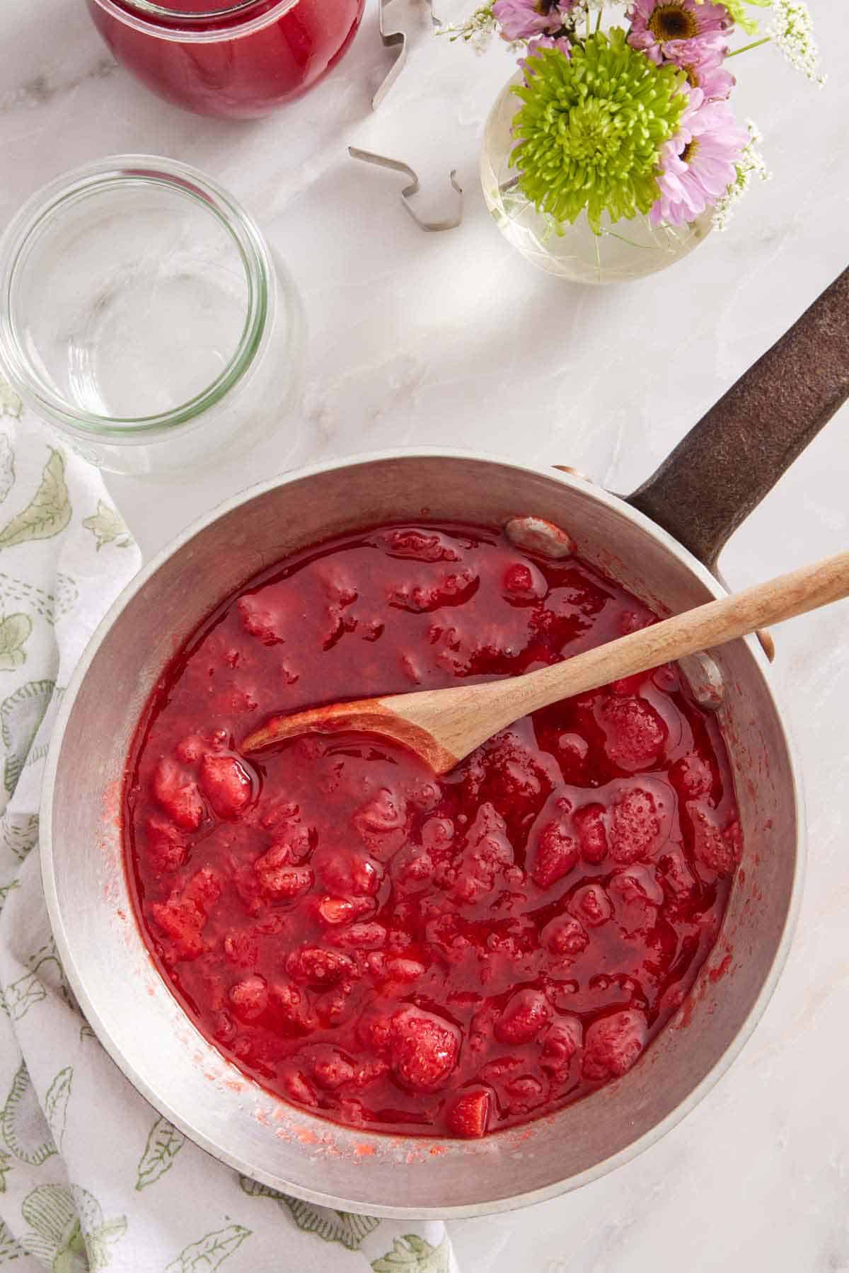 Overhead view of a pot of strawberry sauce with a wooden spoon. Flowers and an empty jar beside it.