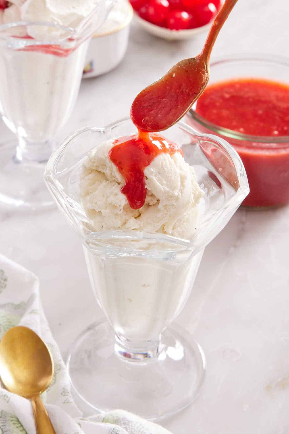 A spoonful of strawberry topping spooned over a tall glass of ice cream. A jar of more sauce in the background and more glasses.