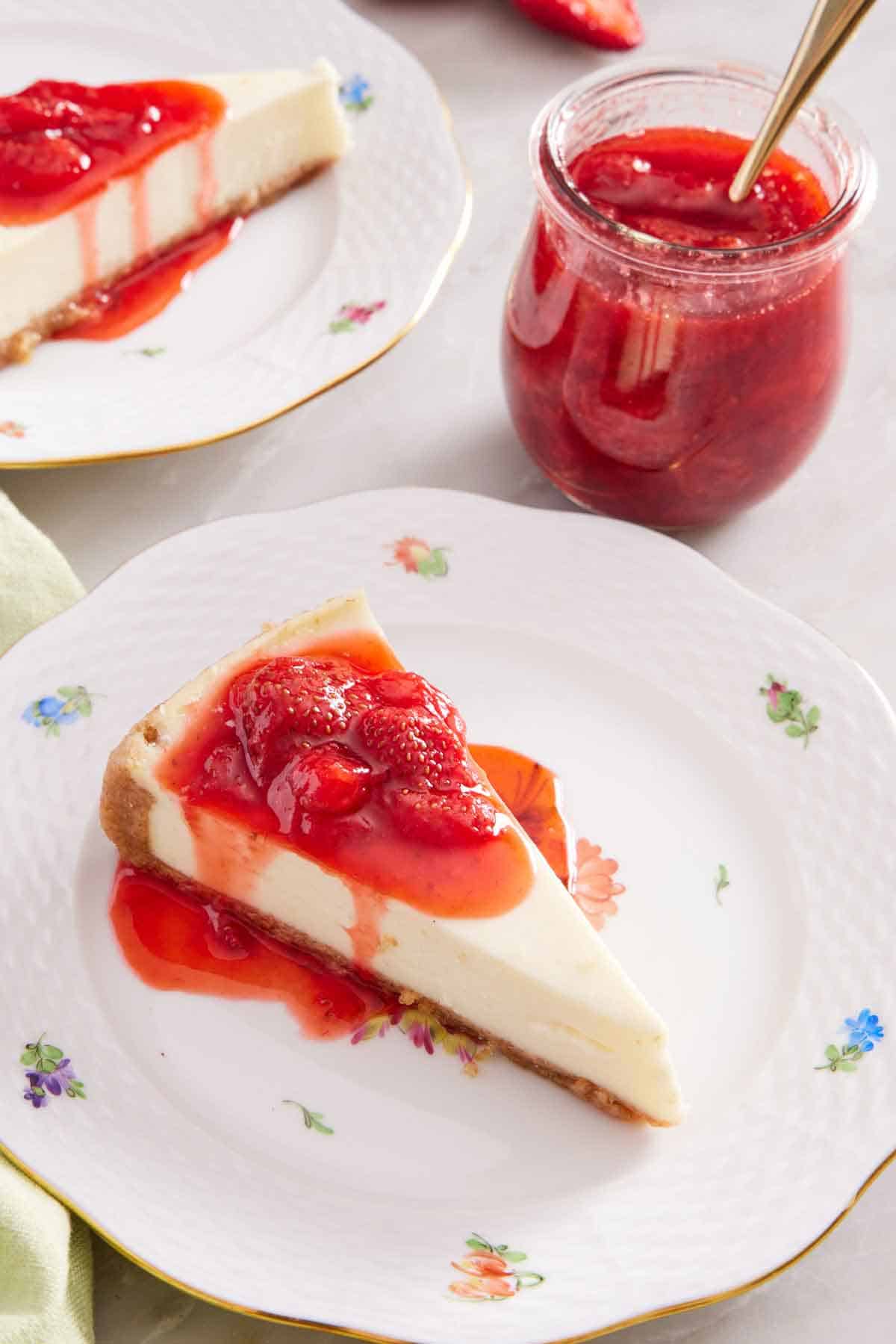 A plate with a slice of cheesecake with strawberry sauce. A jar of more strawberry sauce in the background along with another slice of cheesecake.