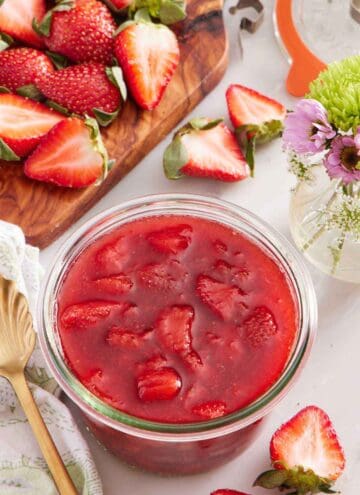 A jar of strawberry sauce. Cut strawberries scattered around and on a cutting board along with a vase of flowers.