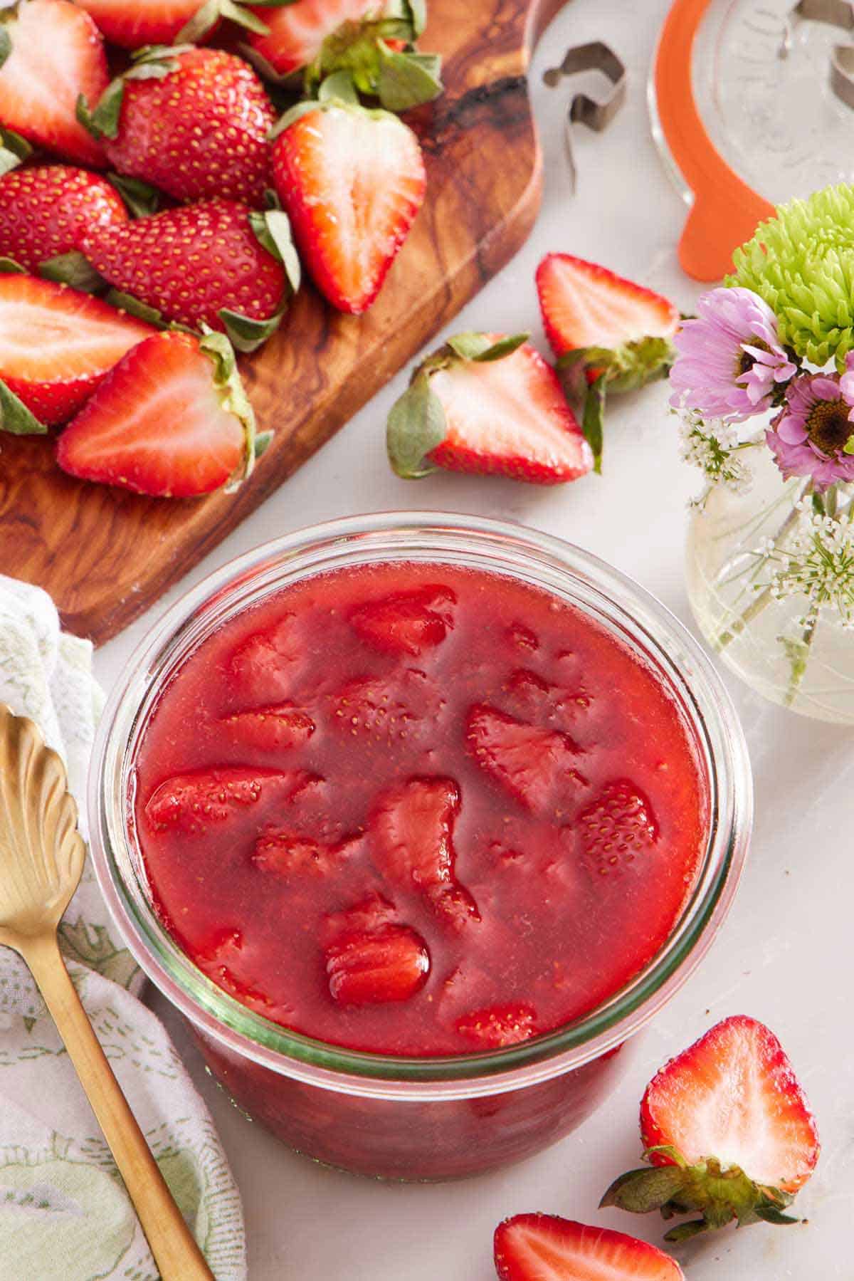 A jar of strawberry sauce. Cut strawberries scattered around and on a cutting board along with a vase of flowers.