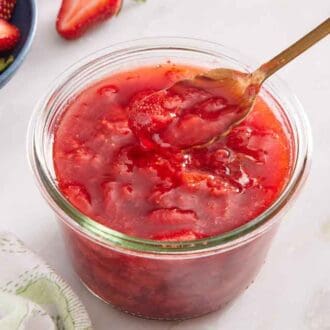 A glass of strawberry sauce with a spoon lifting up a bit of the sauce. Flowers and cut strawberries on the side.