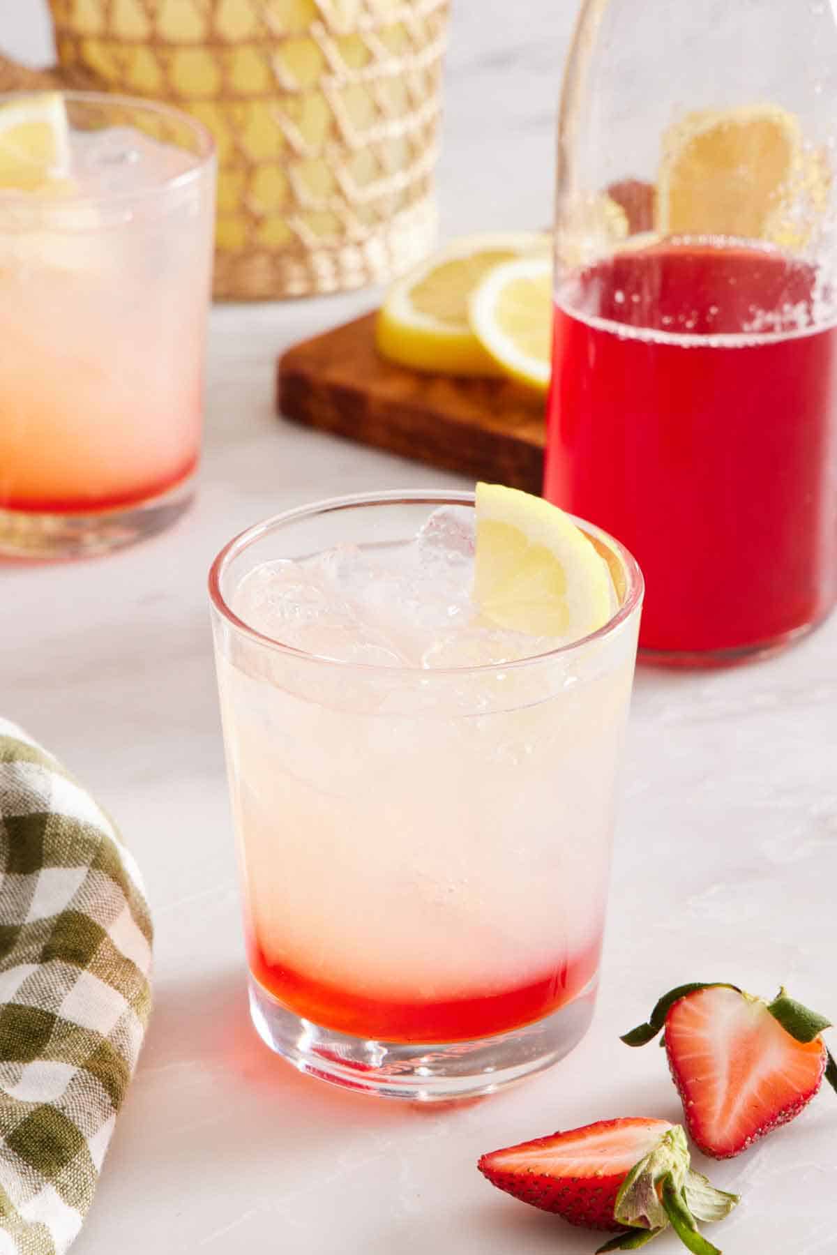 A drink with strawberry syrup on the bottom. A bottle of strawberry syrup in the background along with another drink and some cut strawberries and lemon.