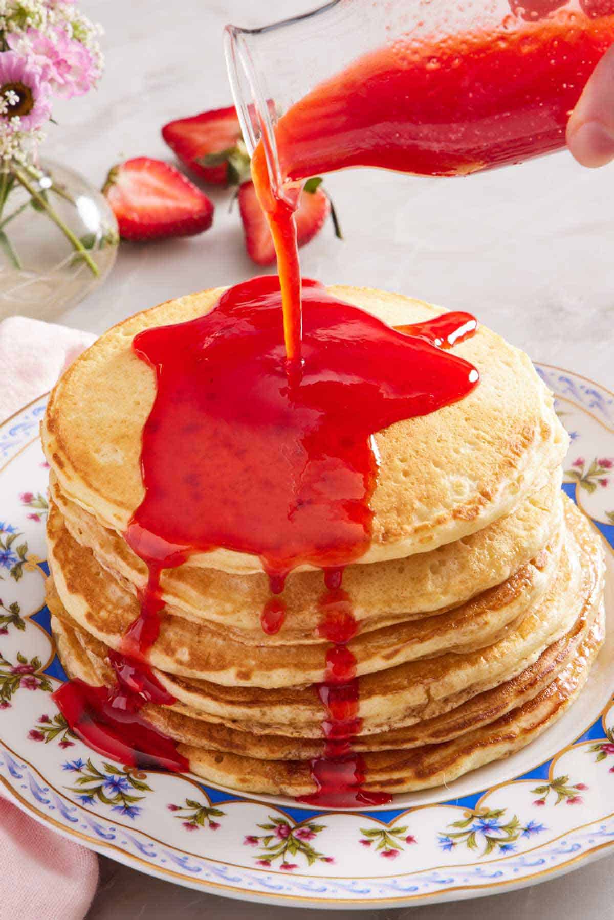 A bottle of strawberry syrup poured over a stack of pancakes on a plate.