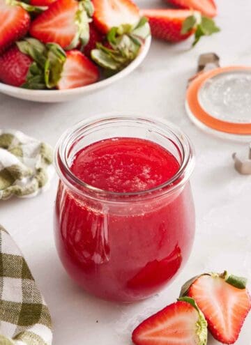 A small glass jar of strawberry syrup. Cut strawberries scattered around and some in a bowl in the background.