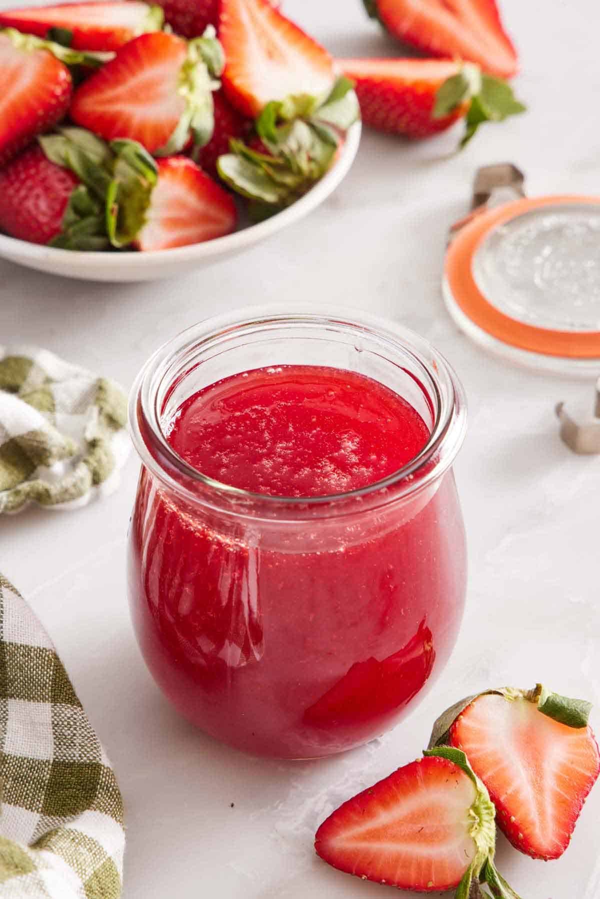 A small glass jar of strawberry syrup. Cut strawberries scattered around and some in a bowl in the background.
