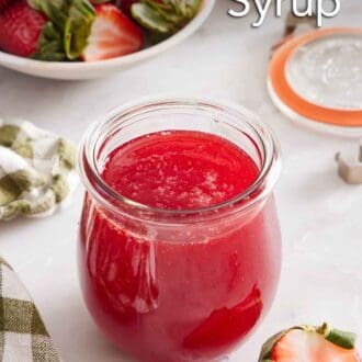 Pinterest graphic of a jar of strawberry syrup. Cut strawberries scattered around and some in a bowl in the background.