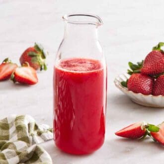 A bottle of strawberry syrup. Strawberries in a bowl and cut strawberries scattered around with a vase of flowers.