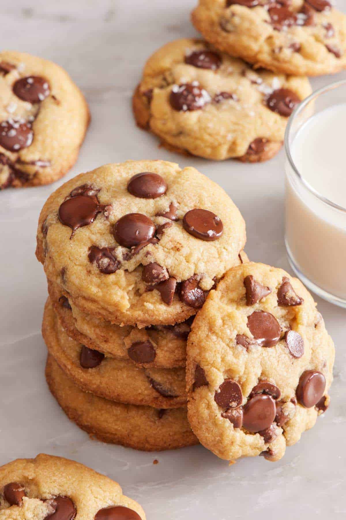 A stack of brown butter chocolate chip cookies with more cookies surrounding it. A glass of milk is on the side.