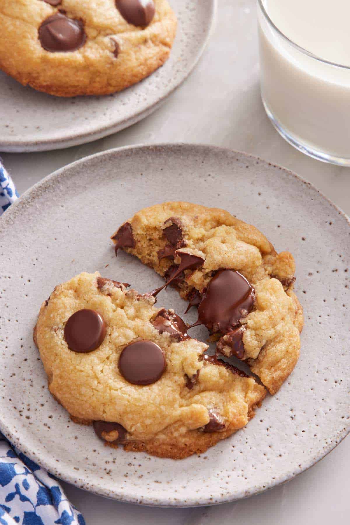 A brown butter cookie with chocolate chips split in half on a plate to showcase the gooey chocolate chips. A glass of milk and another plate with a cookie on the side.