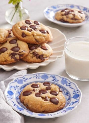 A cookie on a plate next to a glass of milk with more cookies in the background.
