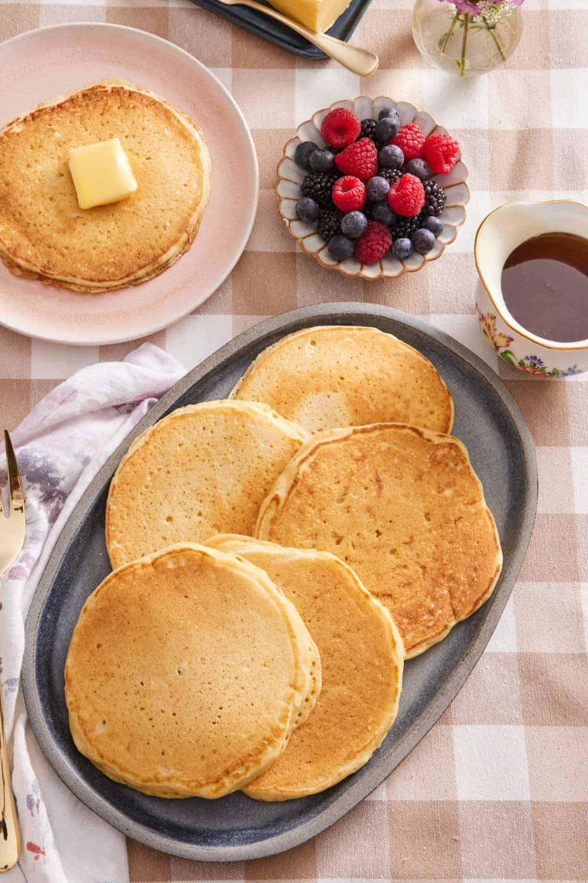 Overhead shot of pancakes on a serving platter, with a single pancake, a bowl of mixed berries, and a small jug of maple syrup on the side.