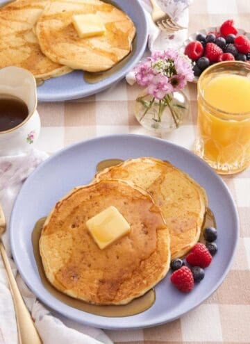 Two buttermilk pancakes on a plate with butter, syrup, and berries. More pancakes, a glass of orange juice, a small vase of flowers, and a bowl of berries in the background.