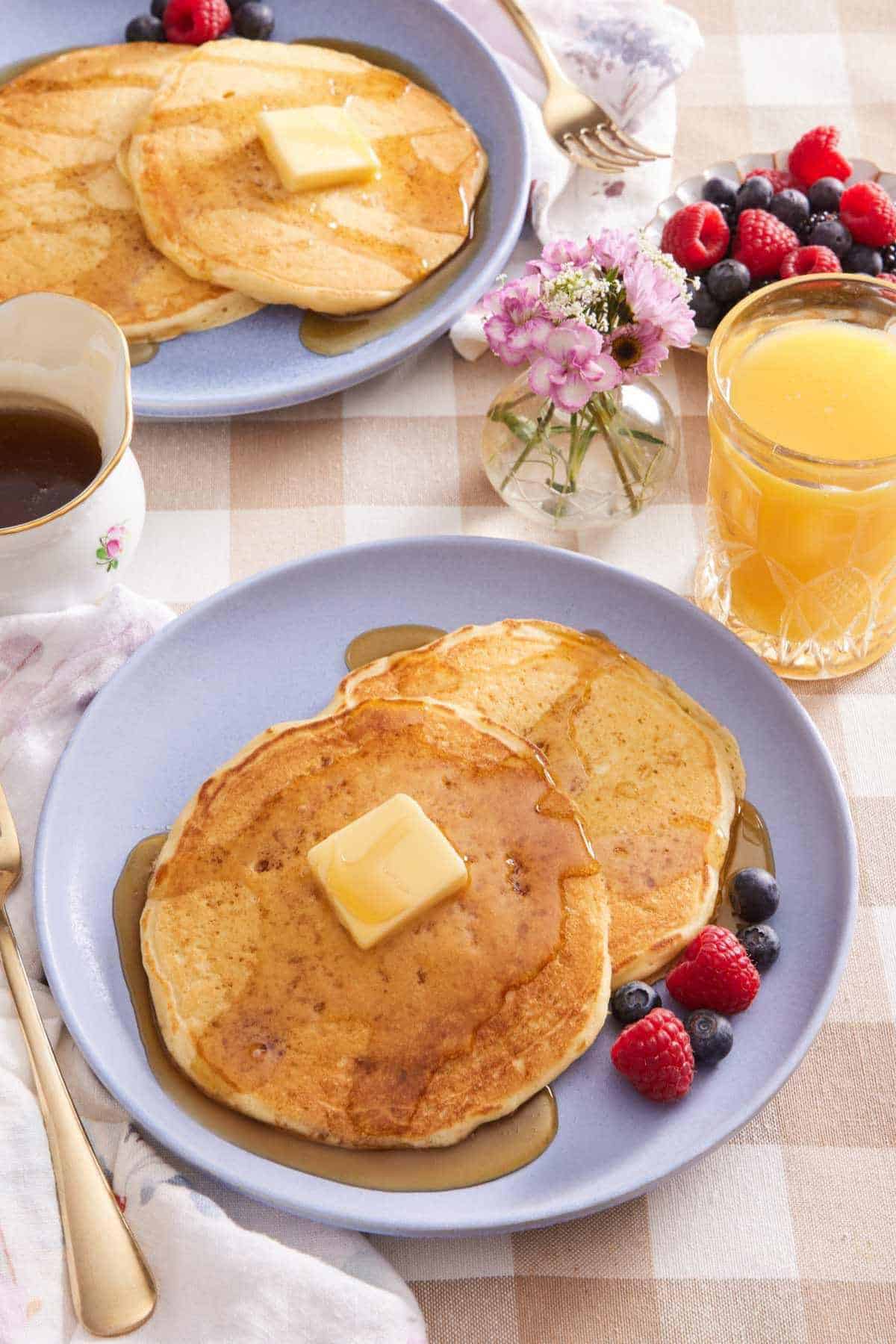Two buttermilk pancakes on a plate with butter, syrup, and berries. More pancakes, a glass of orange juice, a small vase of flowers, and a bowl of berries in the background.