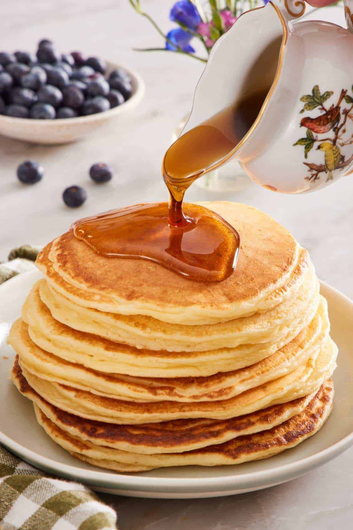 A stack of cottage cheese pancakes on a plate with maple syrup drizzled over the top. A small bowl of blueberries in the background.