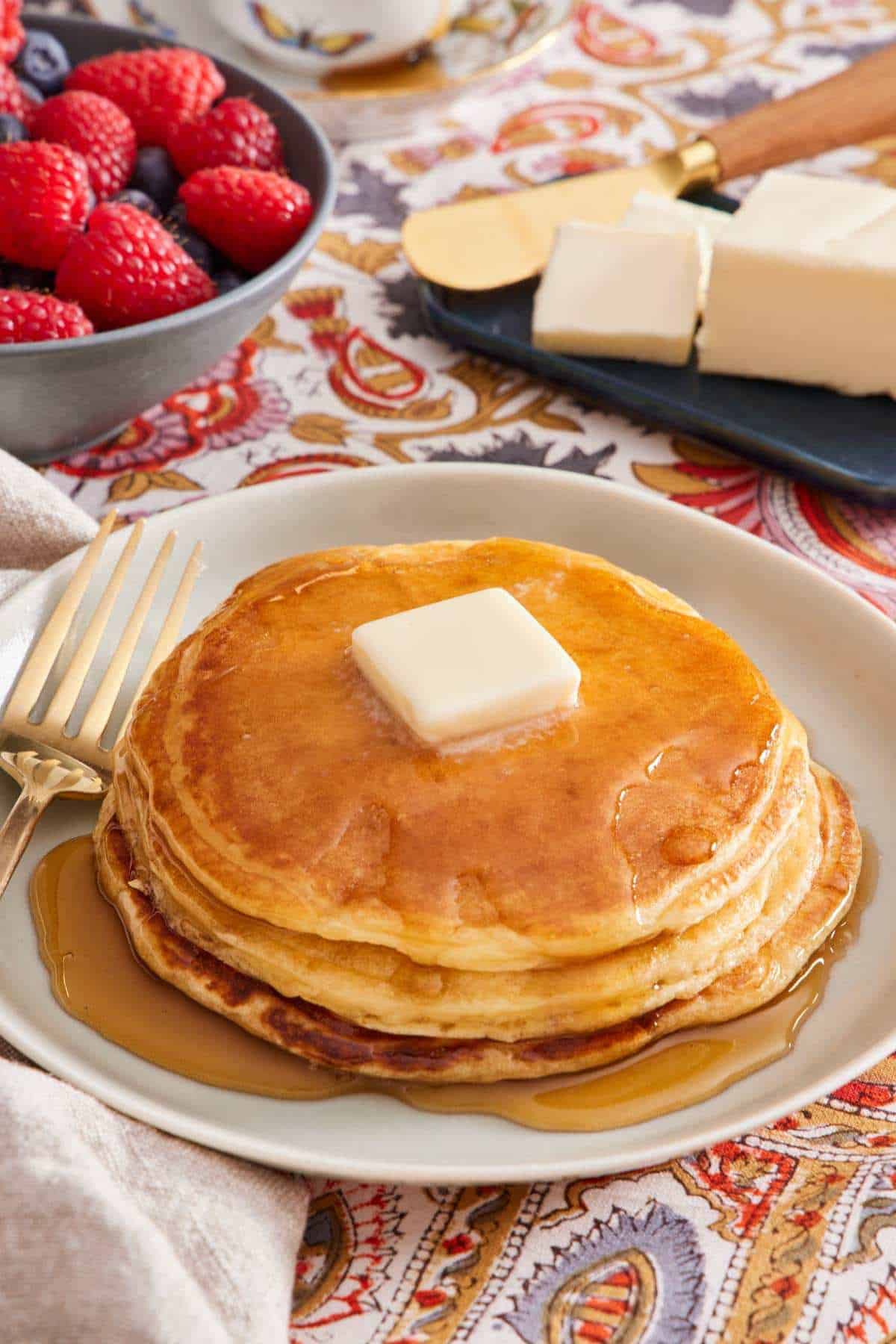 Pancakes with maple syrup and a pat of butter on a plate with mixed berries and a stick of butter in the background.