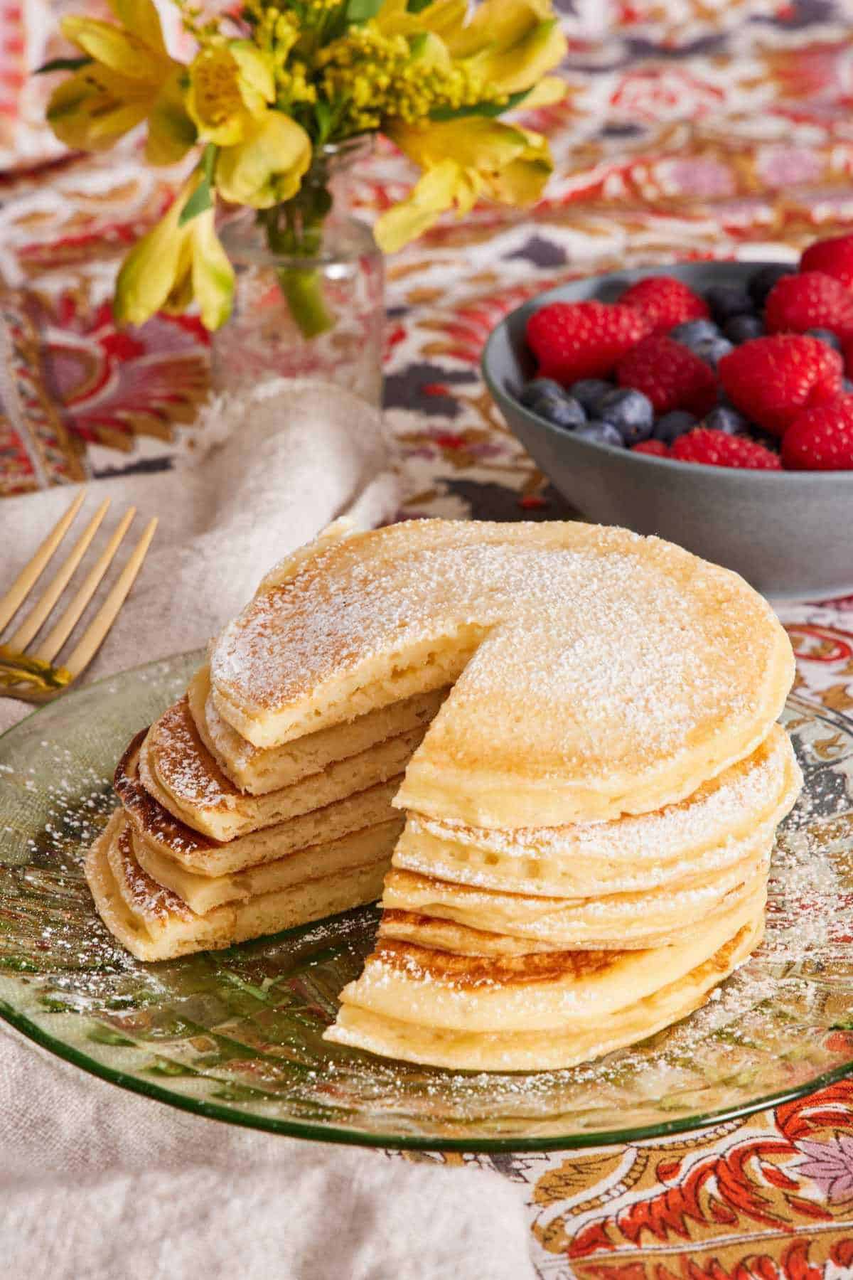 A tall stack of cottage cheese pancakes dusted with powdered sugar. A bowl of mixed berries in the background.