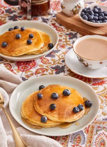 Two plates with pancakes on them, with maple syrup and blueberries on top. A cup of tea is on the side, and more berries in a bowl in the background.