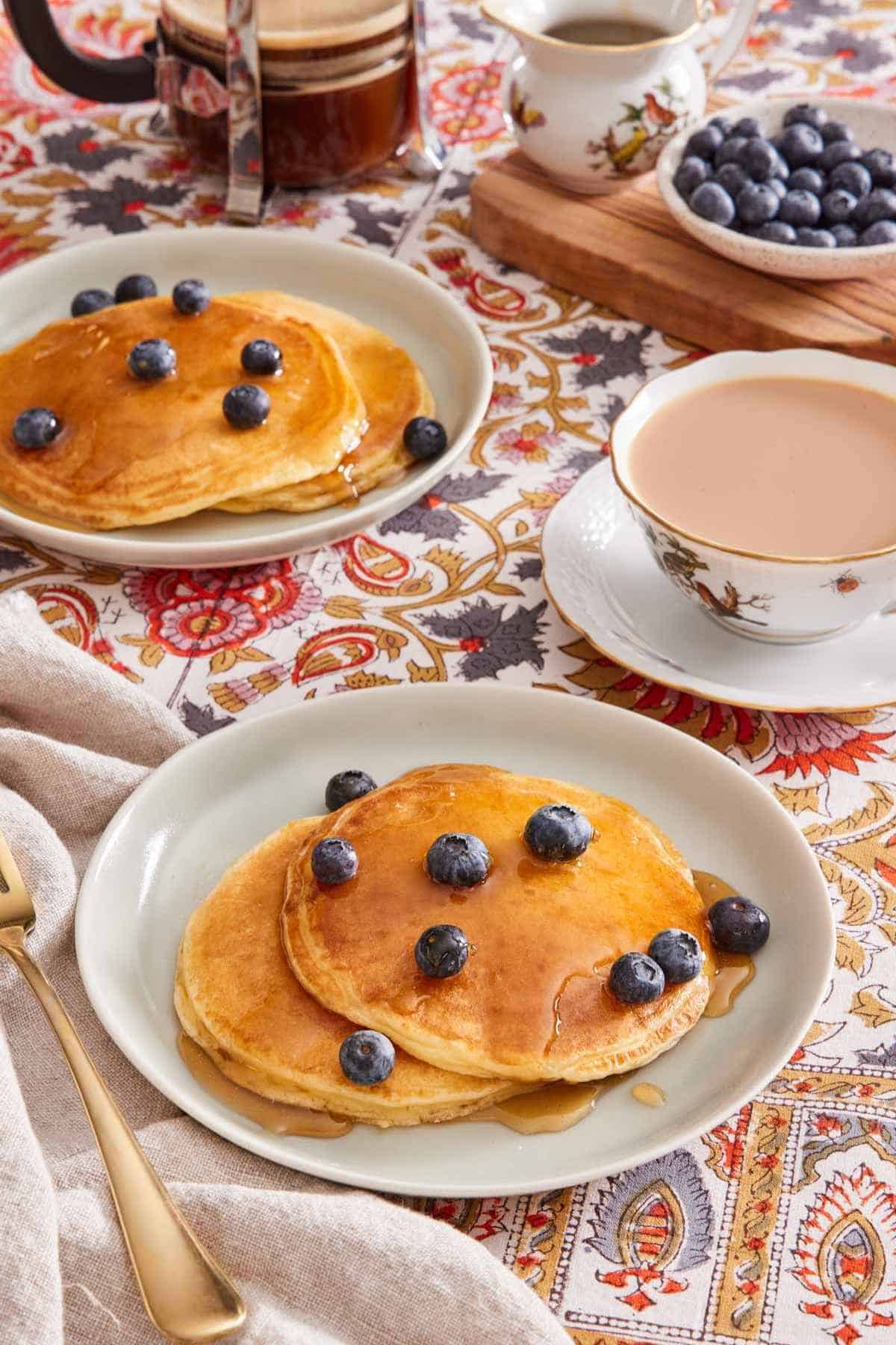 Two plates with pancakes on them, with maple syrup and blueberries on top. A cup of tea is on the side, and more berries in a bowl in the background.