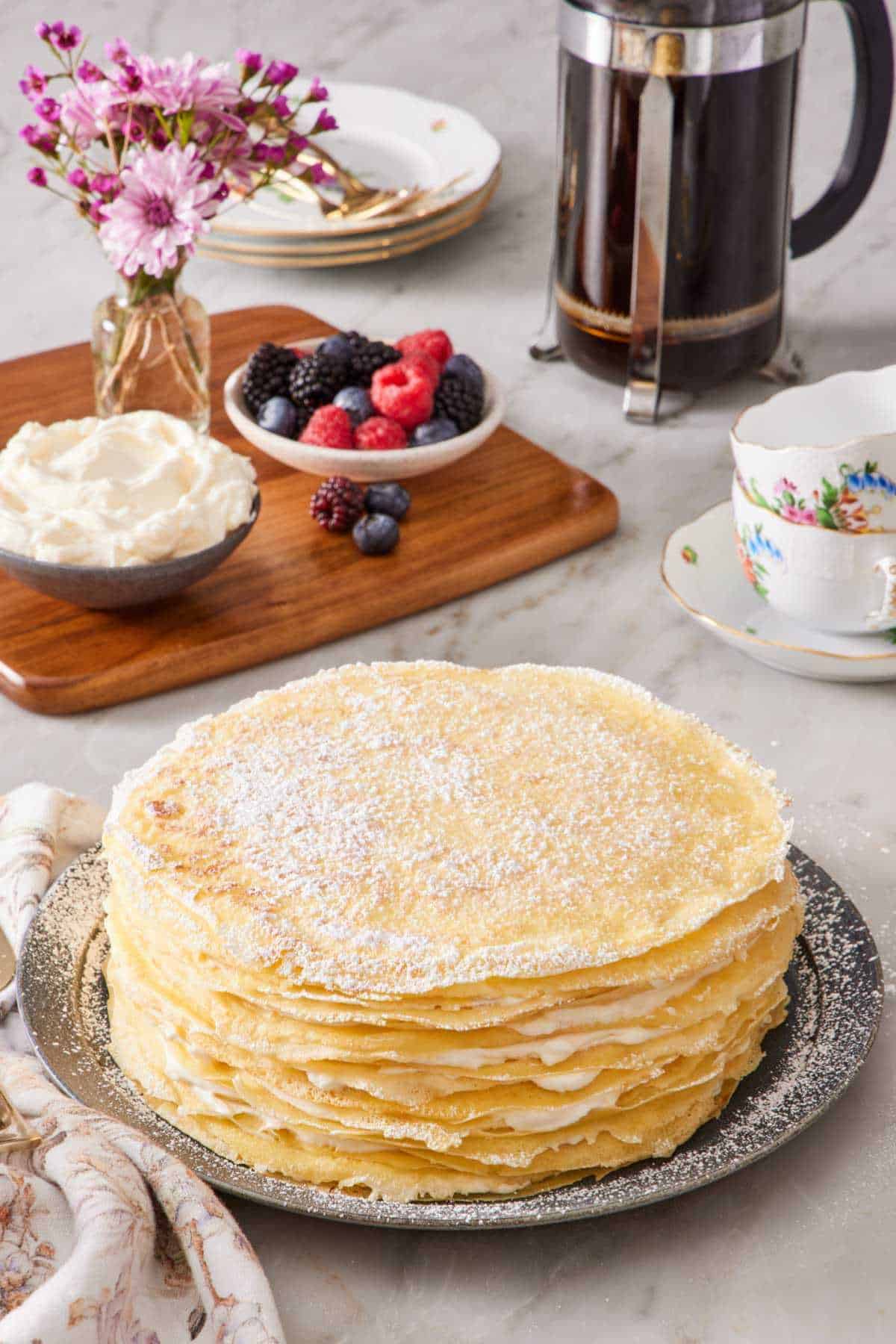 Crepe cake dusted with powdered sugar. A small vase of flowers, a bowl of whipped cream, berries, cups, and a French press with coffee brewing in the background.