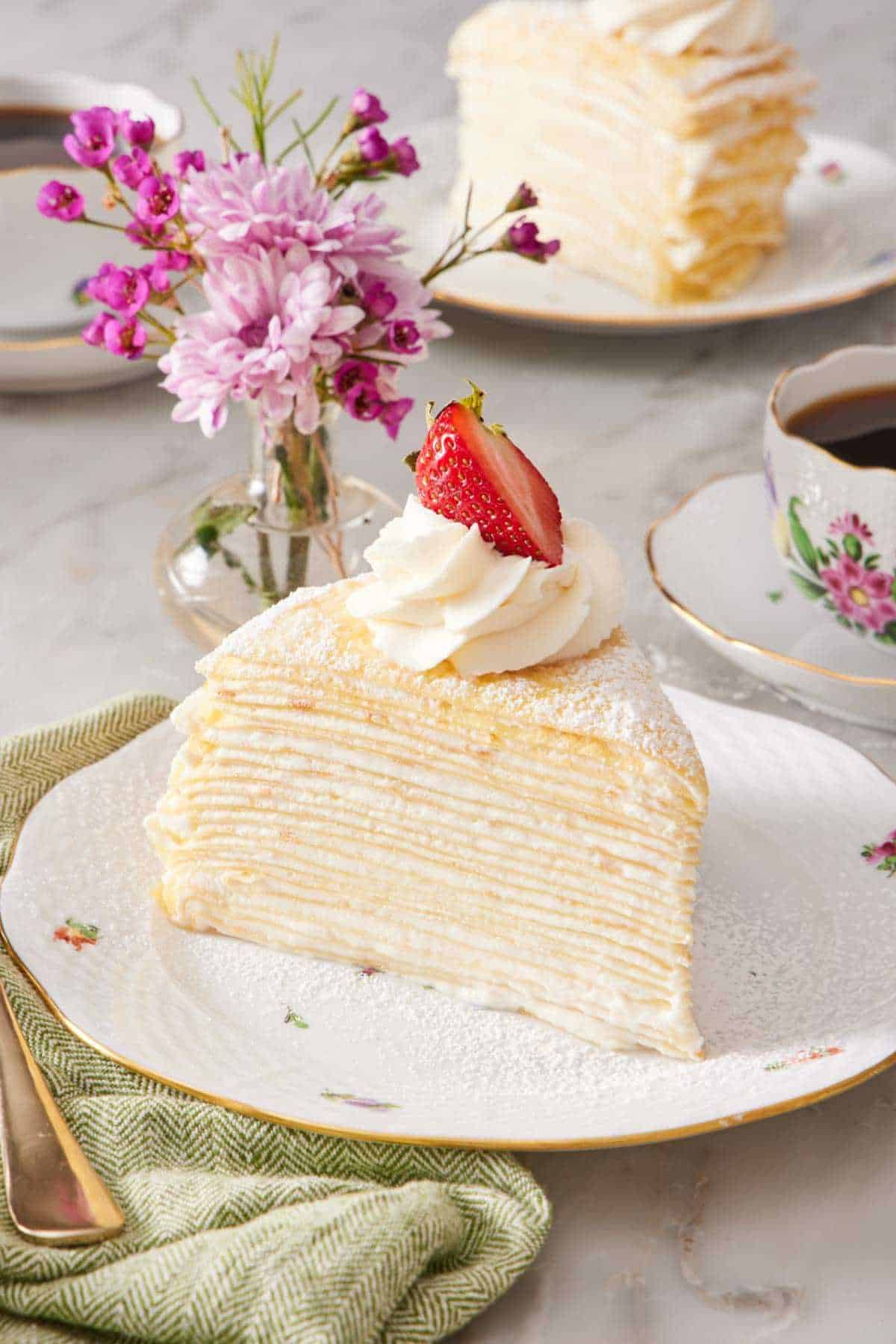 A slice of crepe cake showcasing its many layers. It is garnished with a dollop of whipped cream and halved strawberry. A vase of flowers, a cup of coffee, and another slice of cake are in the background.