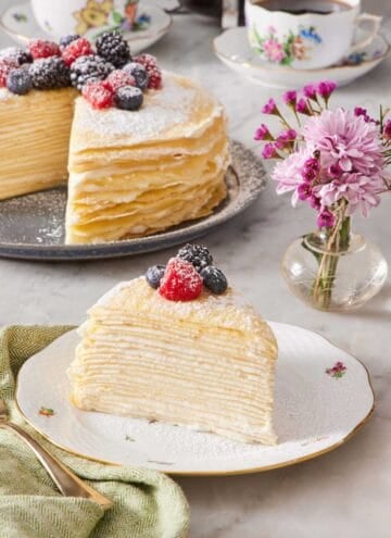 A slice of crepe cake decorated with powdered sugar and berries on a plate. The rest of the unsliced cake, cups of coffee, and a vase of flowers are in the background.