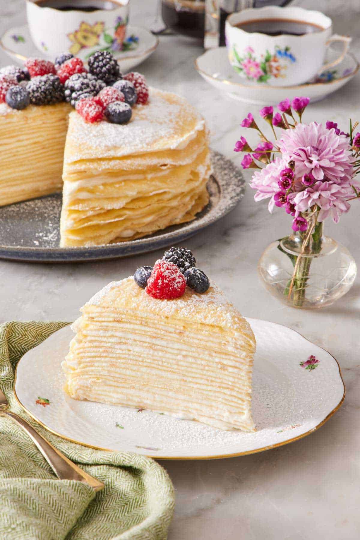 A slice of crepe cake decorated with powdered sugar and berries on a plate. The rest of the unsliced cake, cups of coffee, and a vase of flowers are in the background.