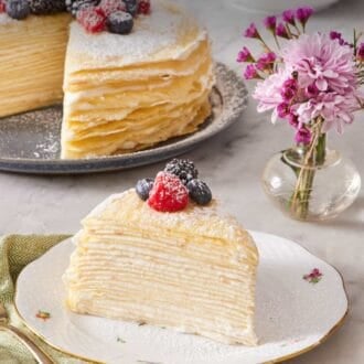 Pinterest graphic of a slice of crepe cake garnished with berries and confectioners' sugar. The rest of the cake, a vase with pink flowers, and cups of coffee are in the background.