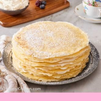 Pinterest graphic showing a crepe cake decorated simply with powdered sugar. In the background, whipped cream and berries are in small bowls on a wooden serving board.