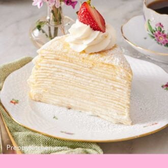 Pinterest graphic of a slice of crepe cake showcasing the delicate crepes and creamy filling. Garnished with a swirl of whipped cream and a halved strawberry. A vase of flowers and cup of coffee are on the side.