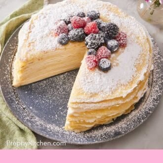 Pinterest graphic of a crepe cake with one slice cut out. Decorated with mixed berries and confectioners' sugar. A bowl of berries and vase of flowers in the background.
