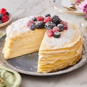 Crepe cake on a serving plate with one slice cut out. It is garnished with fresh berries and powdered sugar. A bowl of berries and a vase of fresh flowers are on the side.