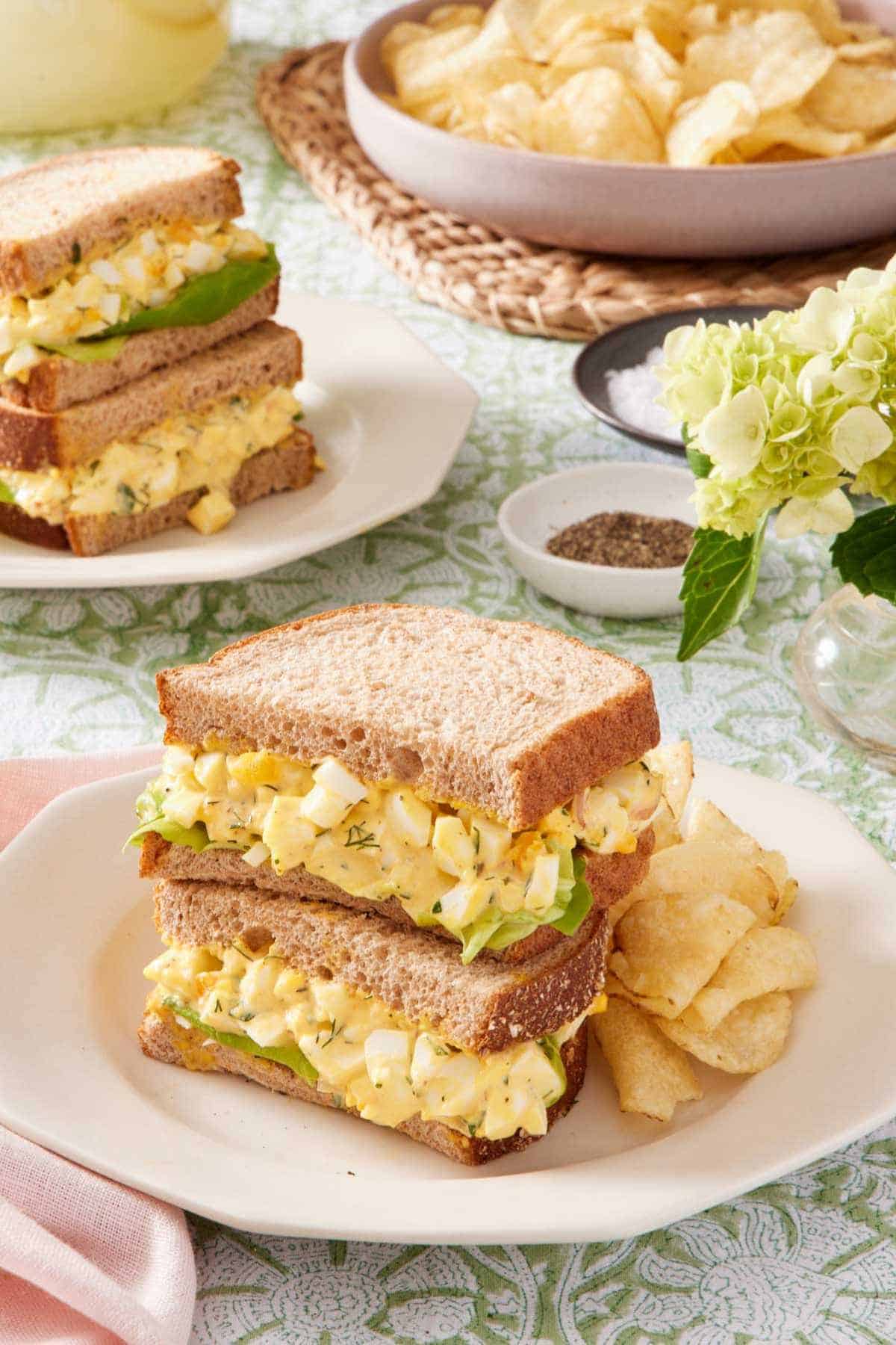Egg salad sandwich and chips on a plate, with another sandwich in the background.