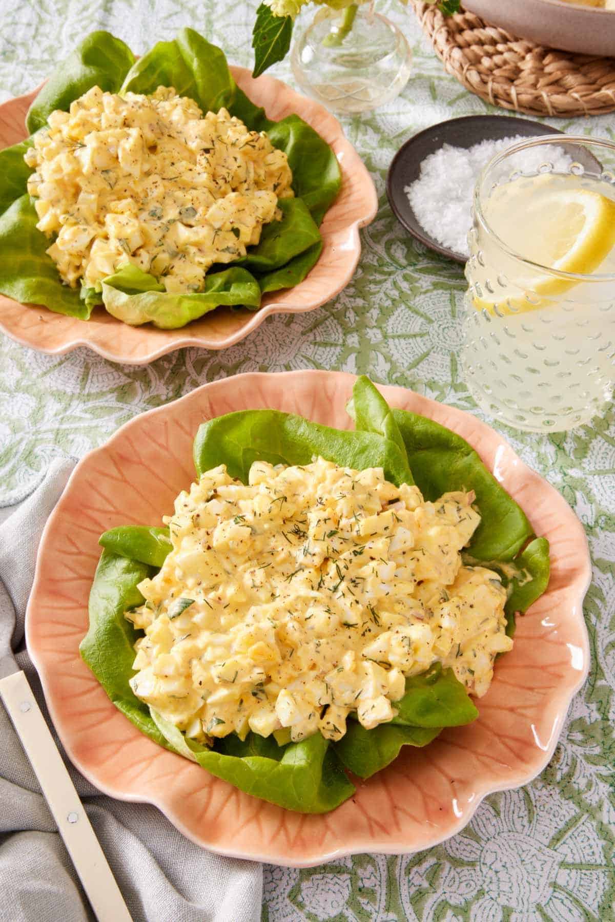 Egg salad served over lettuce on a pink plate. A beverage and another serving plate with salad in the background.