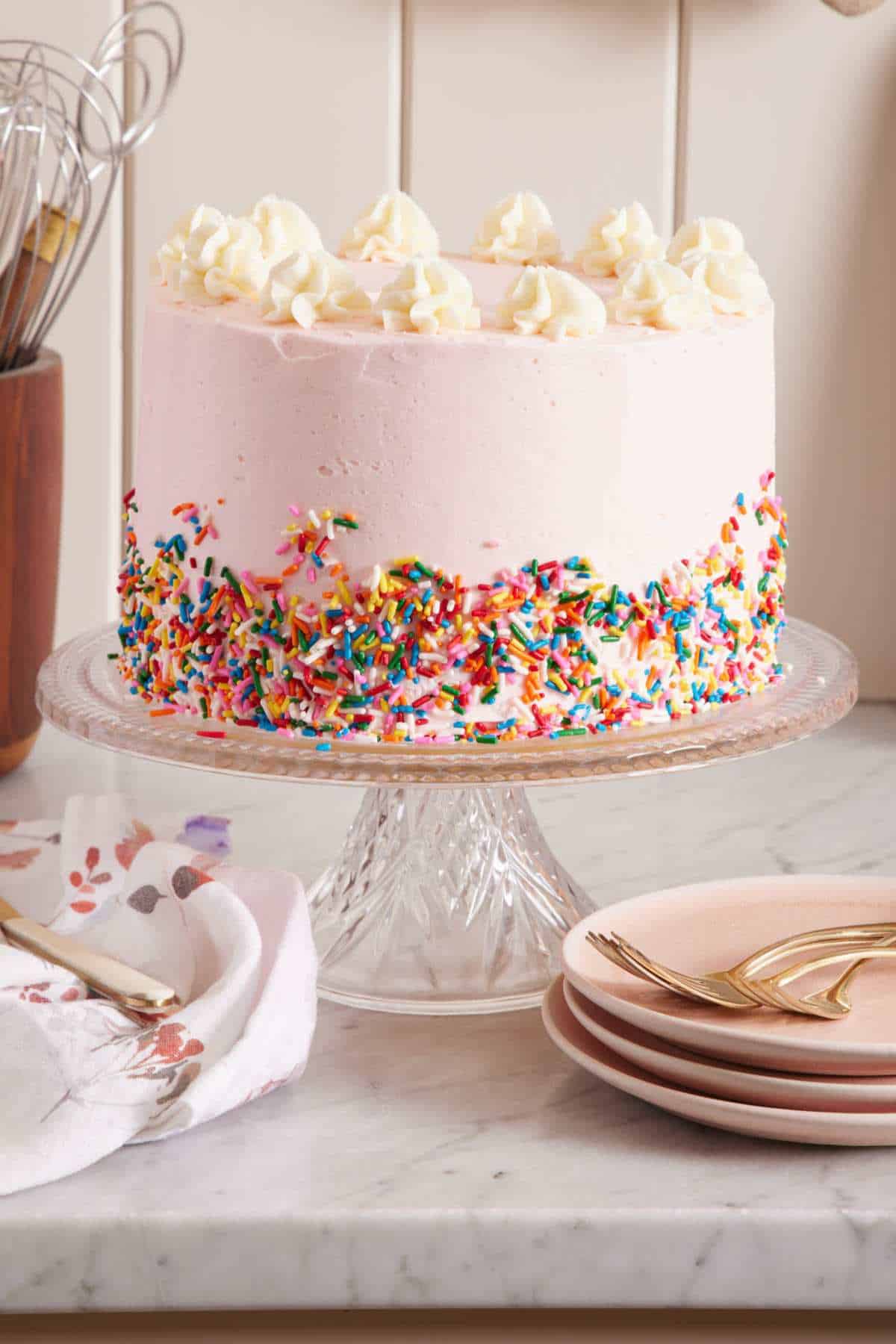 Cake stand holding a layer cake with pink frosting, dollops of frosting on top, and colorful sprinkles pressed around the bottom third of the cake.