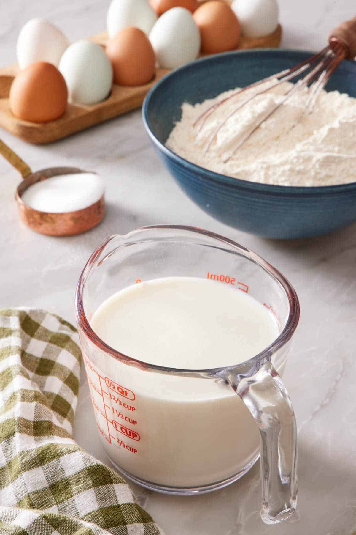 Homemade buttermilk in a measuring cup with flour, sugar, and eggs in the background.