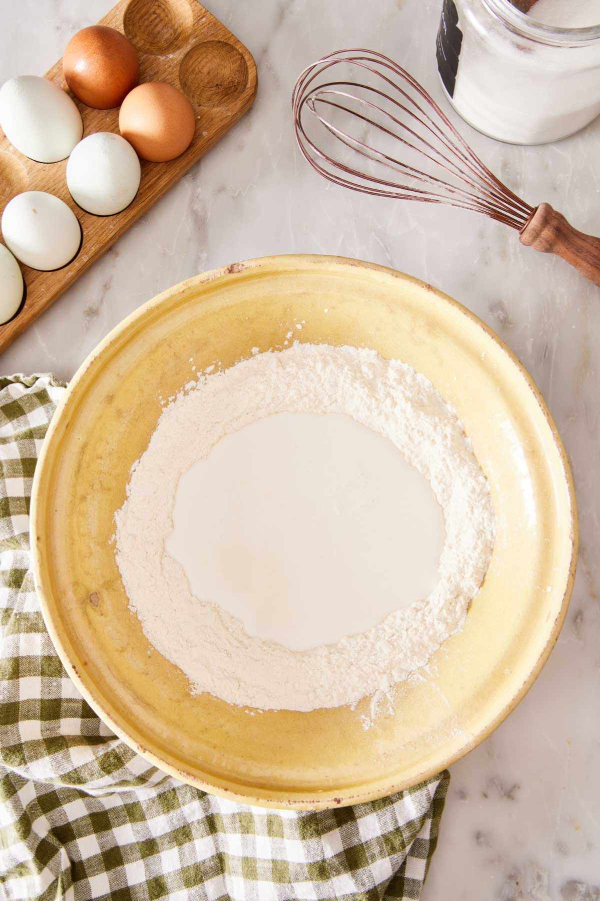 Overhead shot of a bowl with dry ingredients and buttermilk added. A whisk and eggs in a wooden holder in the background.