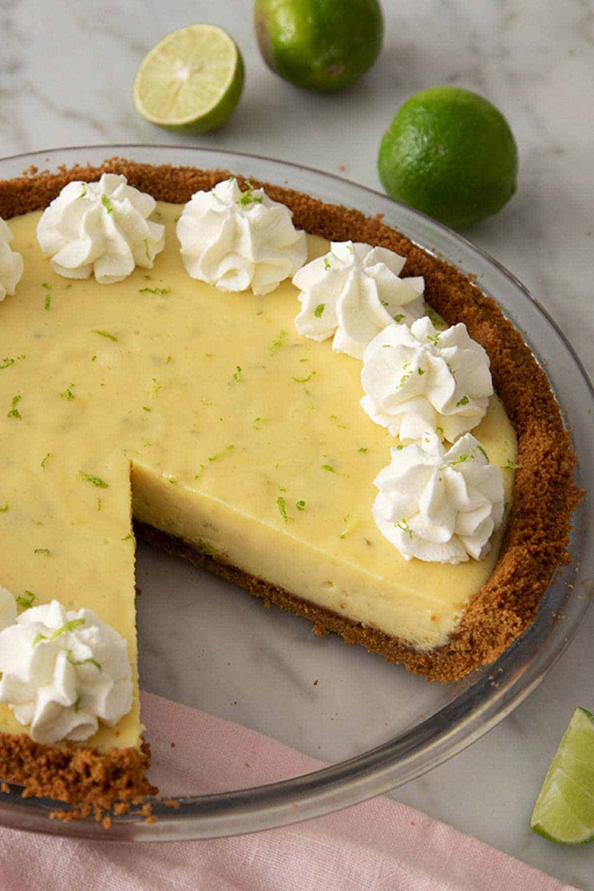 Key lime pie in a pie dish with a slice cut out. Topped with dollops of whipped cream.
