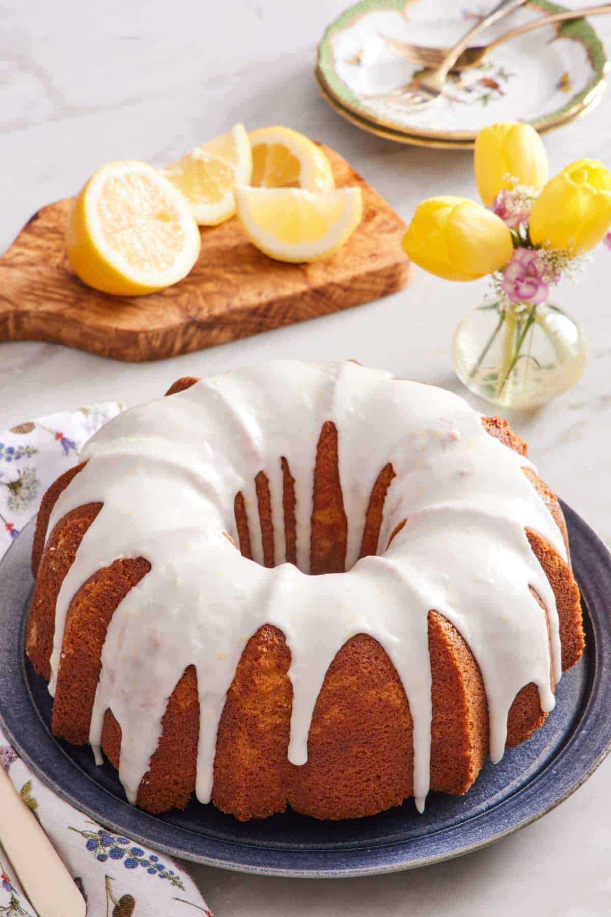 Unsliced lemon bundt cake with powdered sugar glaze. A sliced lemon is on a wooden cutting board in the background, next to a small vase of yellow flowers.