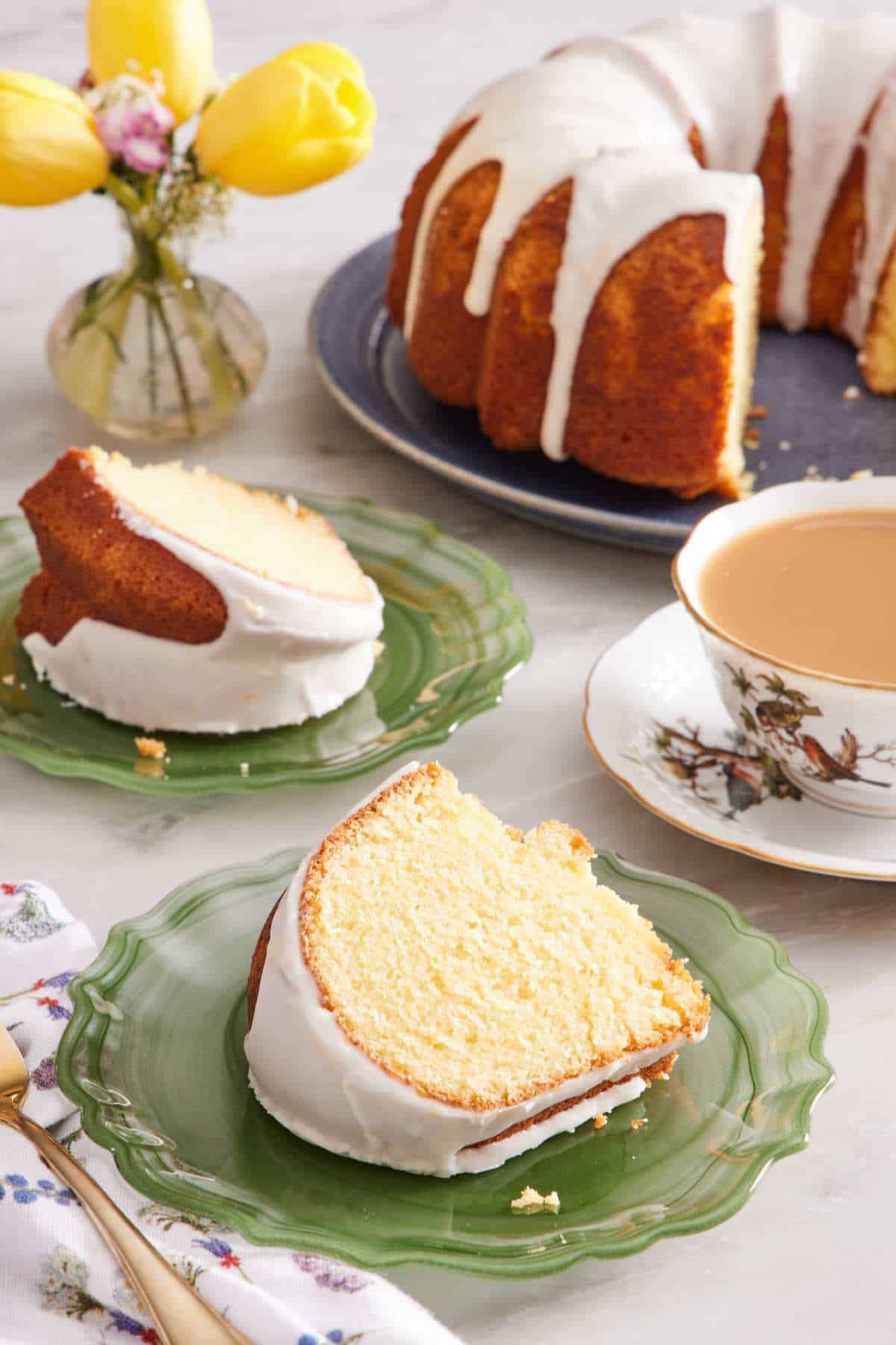 A slice of lemon bundt cake with more cake and a cup of tea in the background.