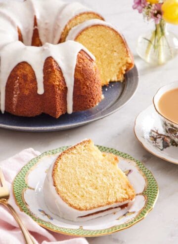 A slice of bundt cake with lemon glaze on a small plate. A cup of tea and the rest of the cake is in the background.