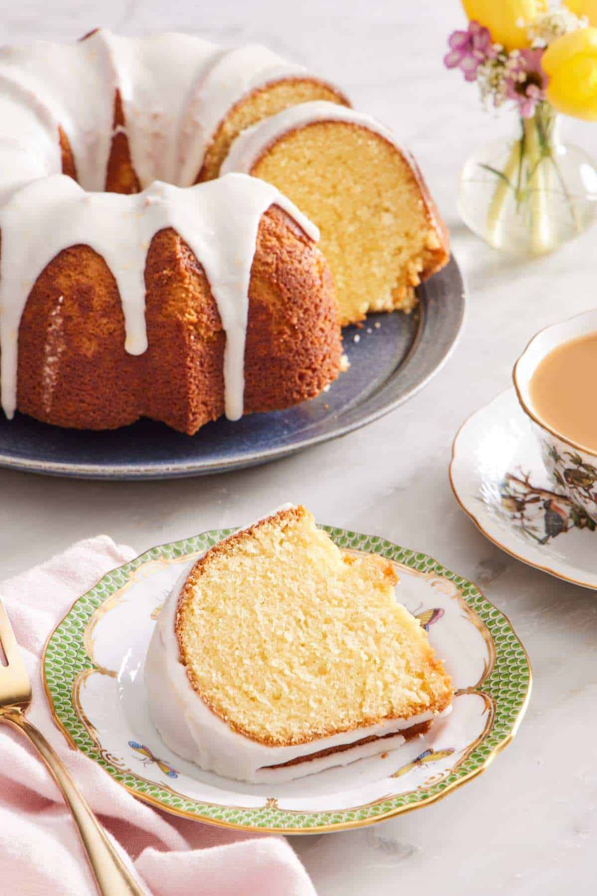 A slice of bundt cake with lemon glaze on a small plate. A cup of tea and the rest of the cake is in the background.