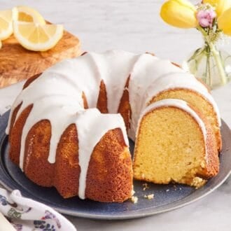 Lemon bundt cake on a cake plate with a few slices cut out. Small serving plates, a cake server, and sliced lemons are in the background.