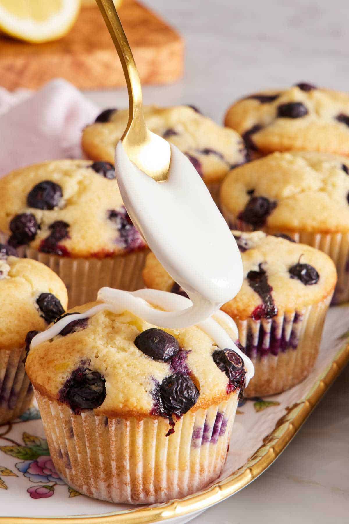 A platter of blueberry muffins and lemon glaze being drizzled on top with a spoon.