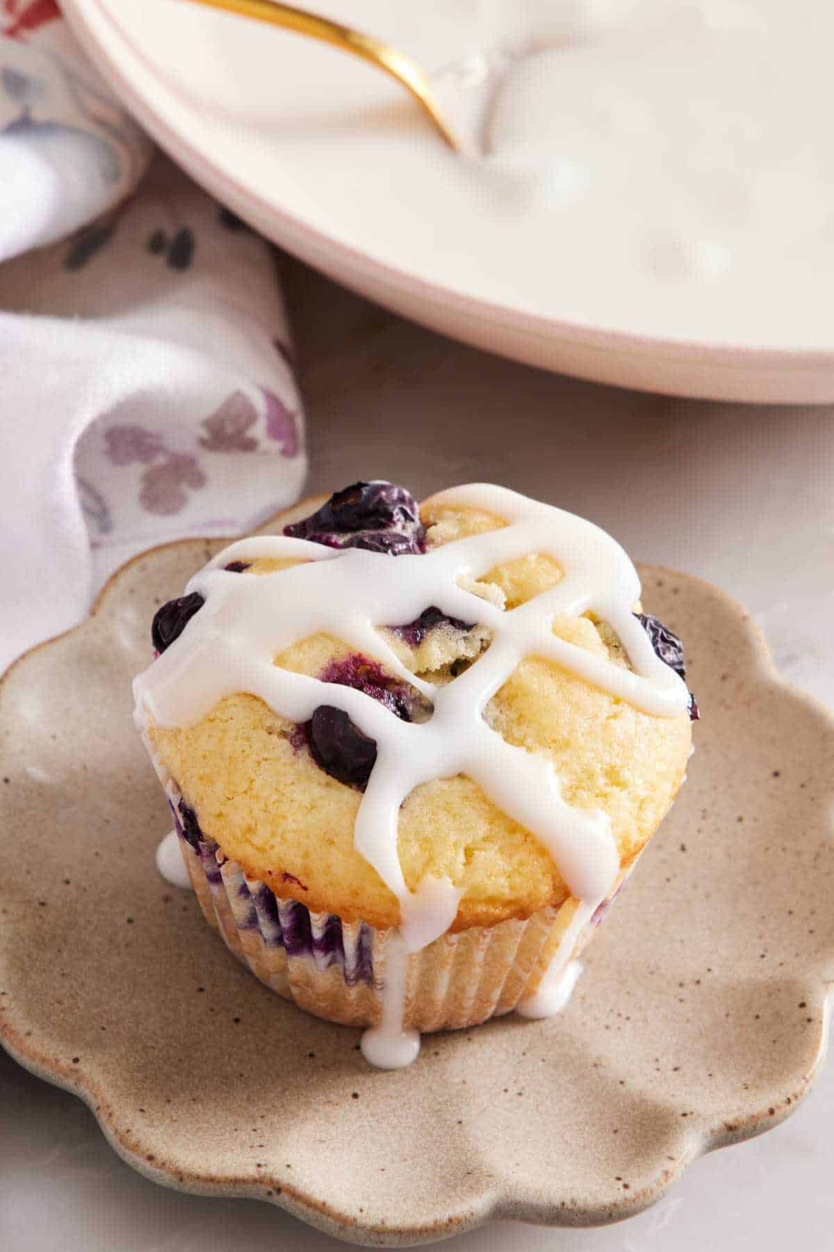 Blueberry muffin on a plate with citrus glaze drizzled on top. A bowl of homemade glaze in the background.
